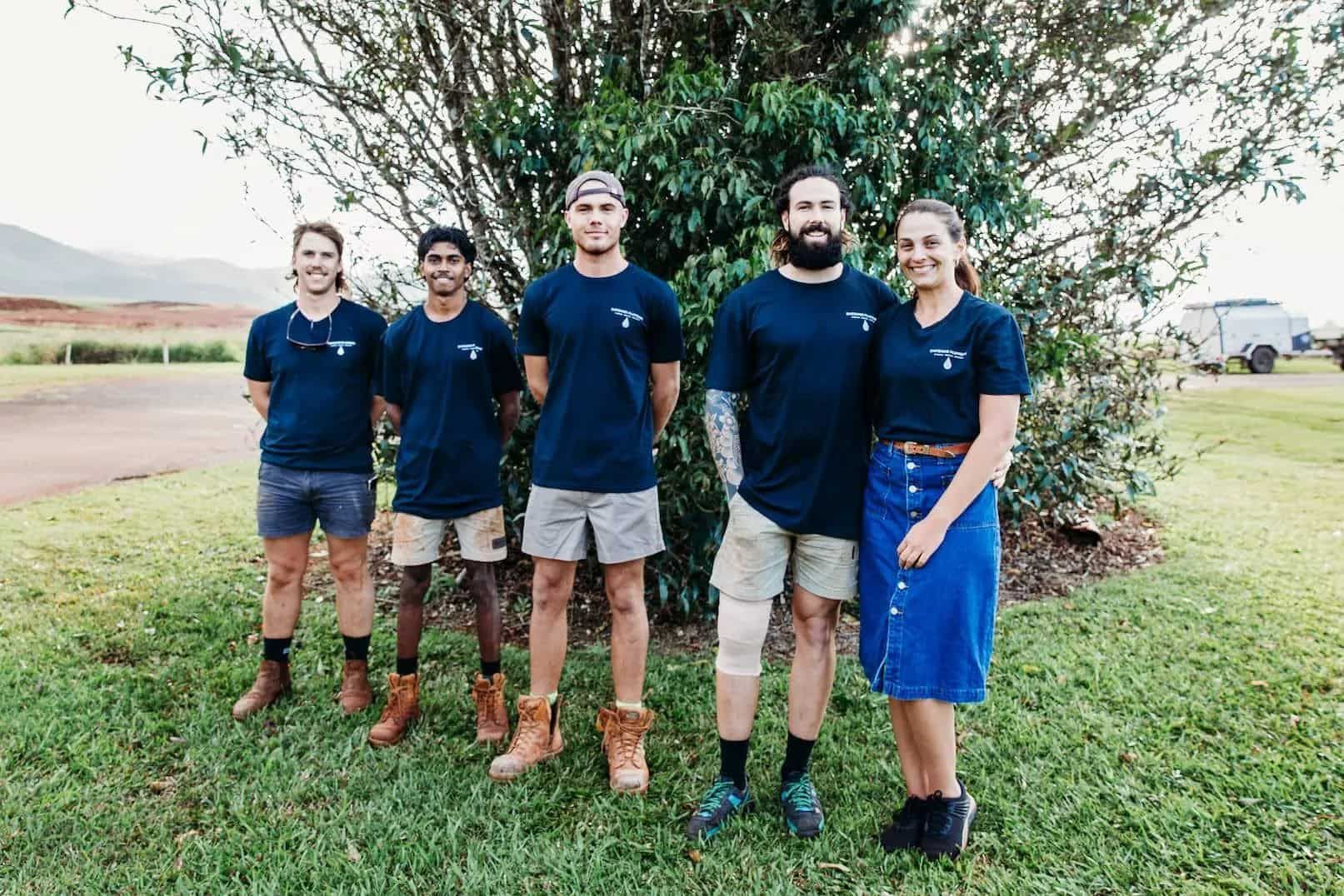 Five people in matching blue shirts pose outside on a grassy area, a tree behind them.