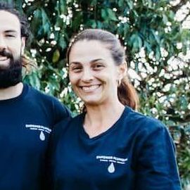 Woman smiling, standing near a man. Both wearing blue shirts, outdoors near foliage.