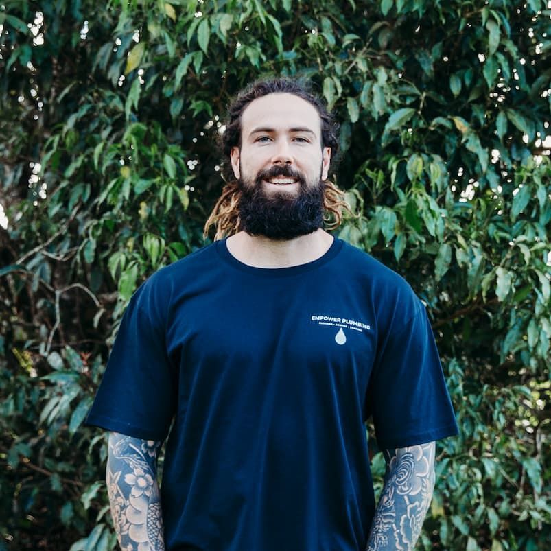 Man with beard and tattoos smiles, wearing a dark blue t-shirt, in front of green foliage.