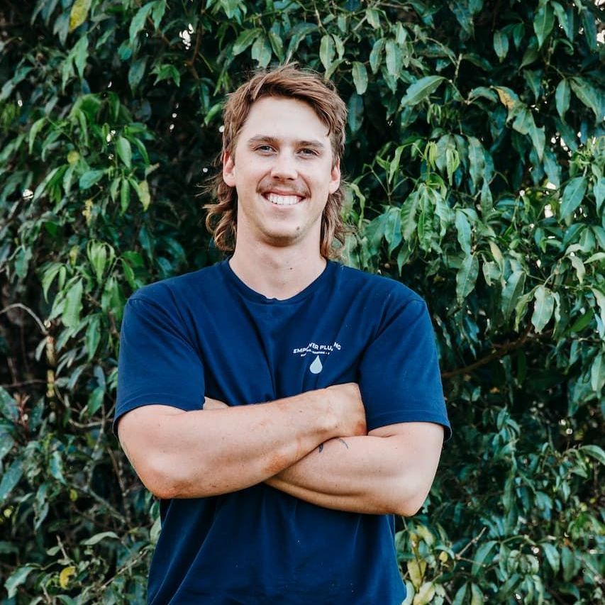 Man with a mullet smiles, arms crossed, in front of leafy green foliage. He wears a dark blue t-shirt.