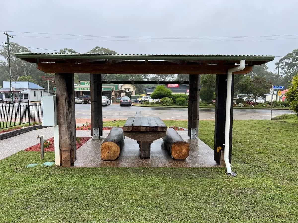 Picnic shelter with wooden table and benches in a grassy area, rain falling. Road and buildings in background.