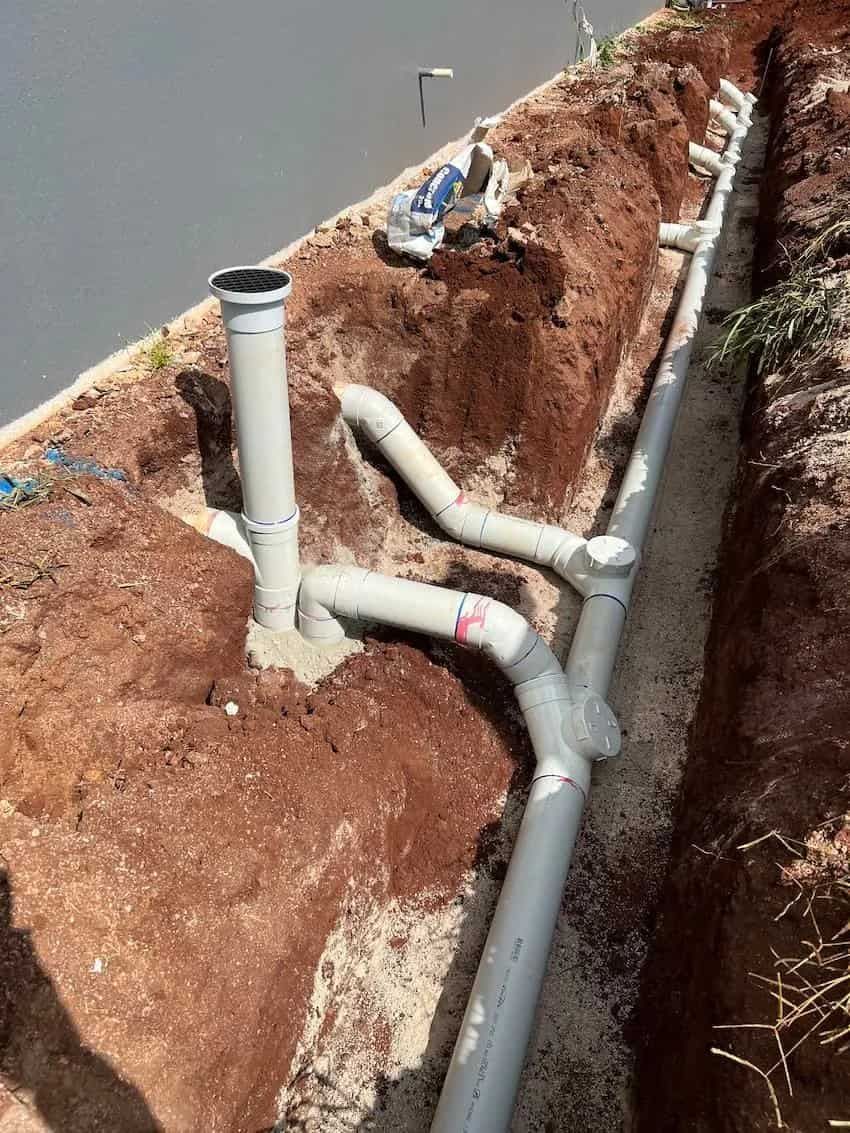 Pipes installed in a trench, near concrete, for drainage. Grey PVC pipes with various fittings; dirt and gravel visible.