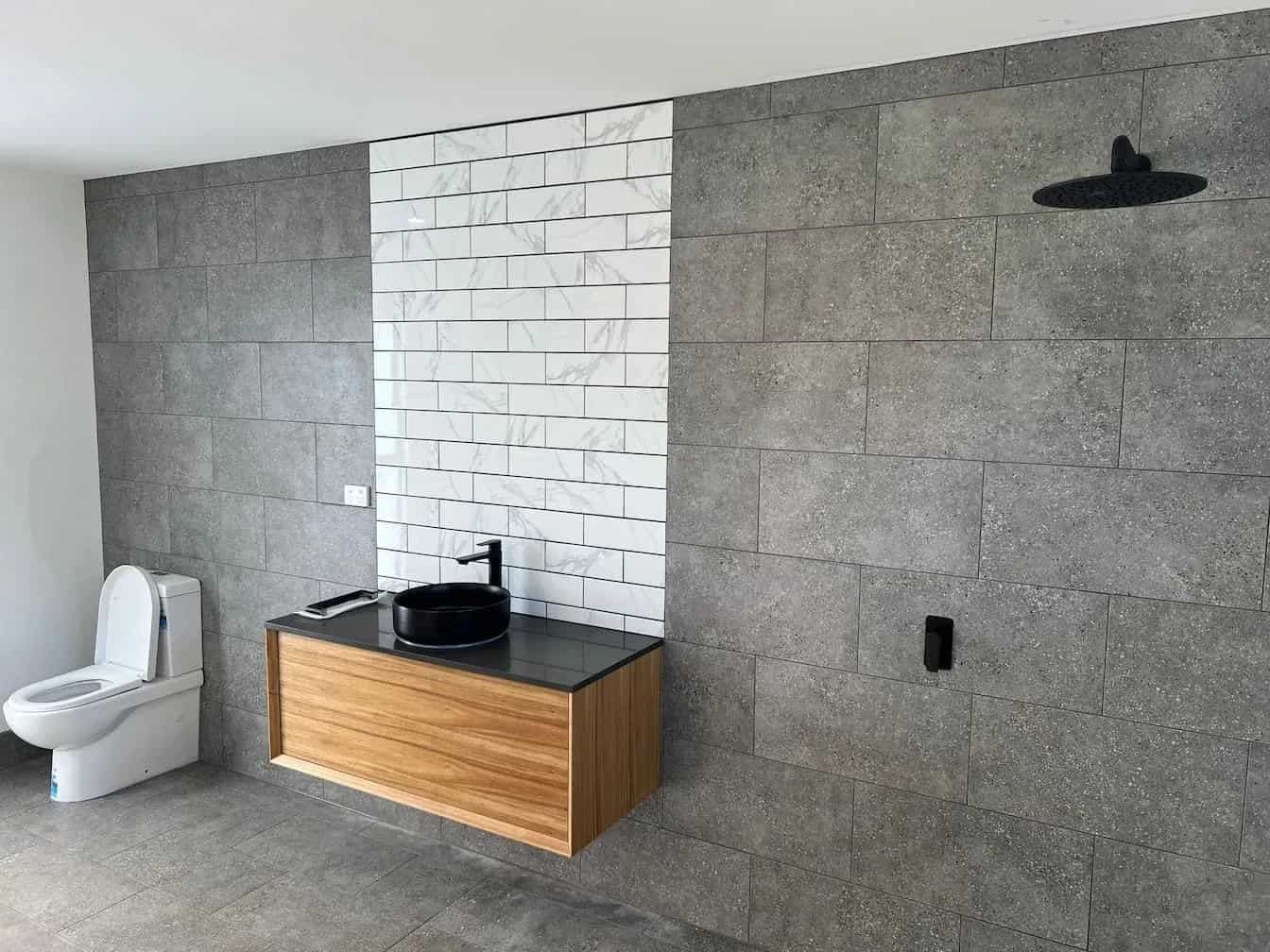 Bathroom with grey stone walls, white toilet, wooden vanity, black sink, and rainfall showerhead.