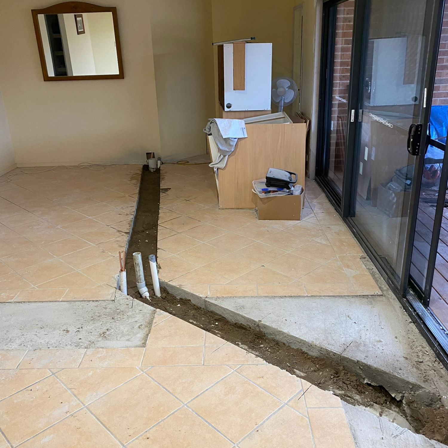 Renovation in a room with broken floor tiles, plumbing exposed, and sliding glass door.