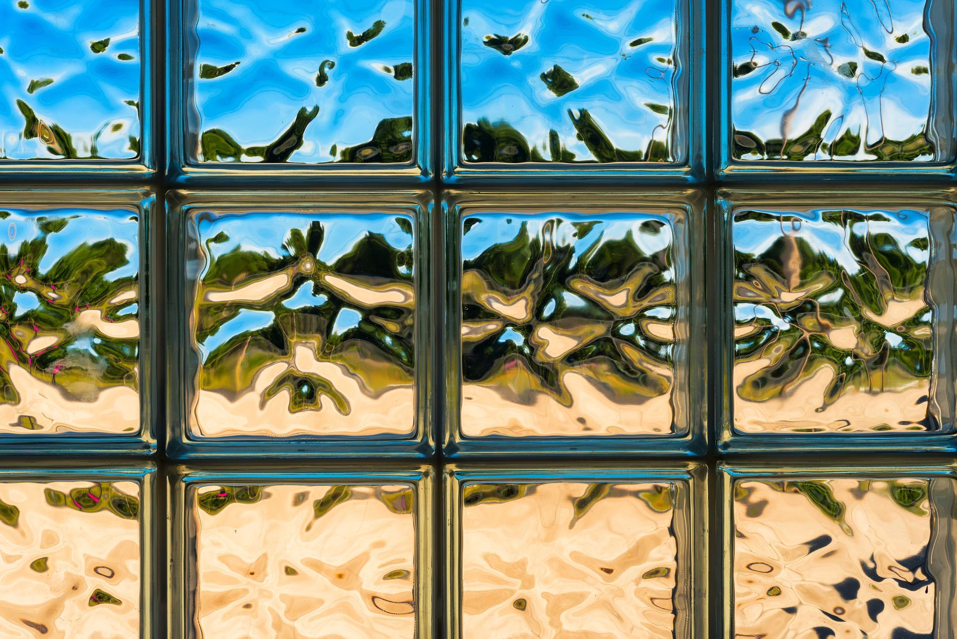 Glass block wall reflecting a blue sky, trees, and a beige surface.