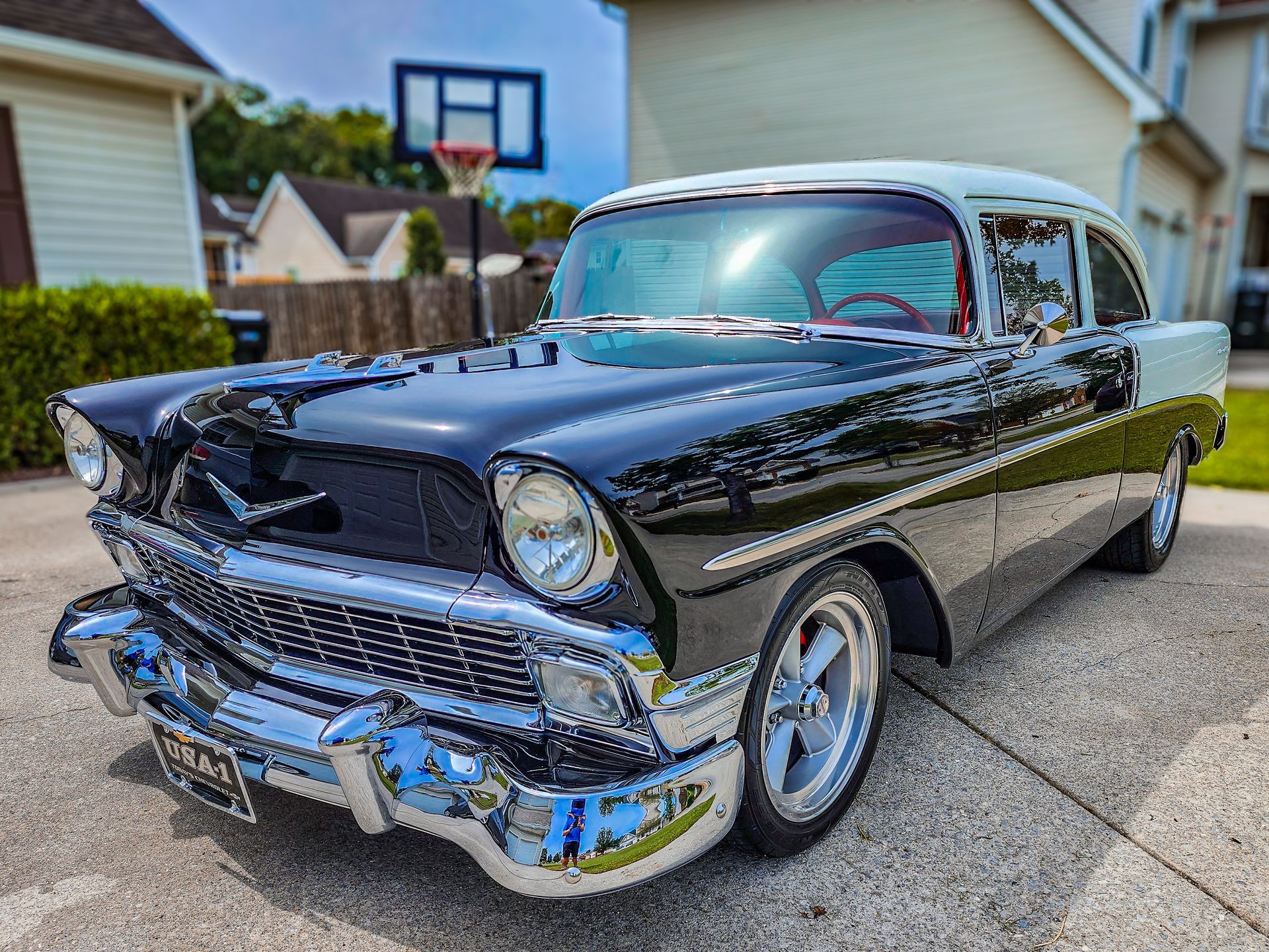 A black and white classic car is parked in front of a house with a basketball hoop in the background.