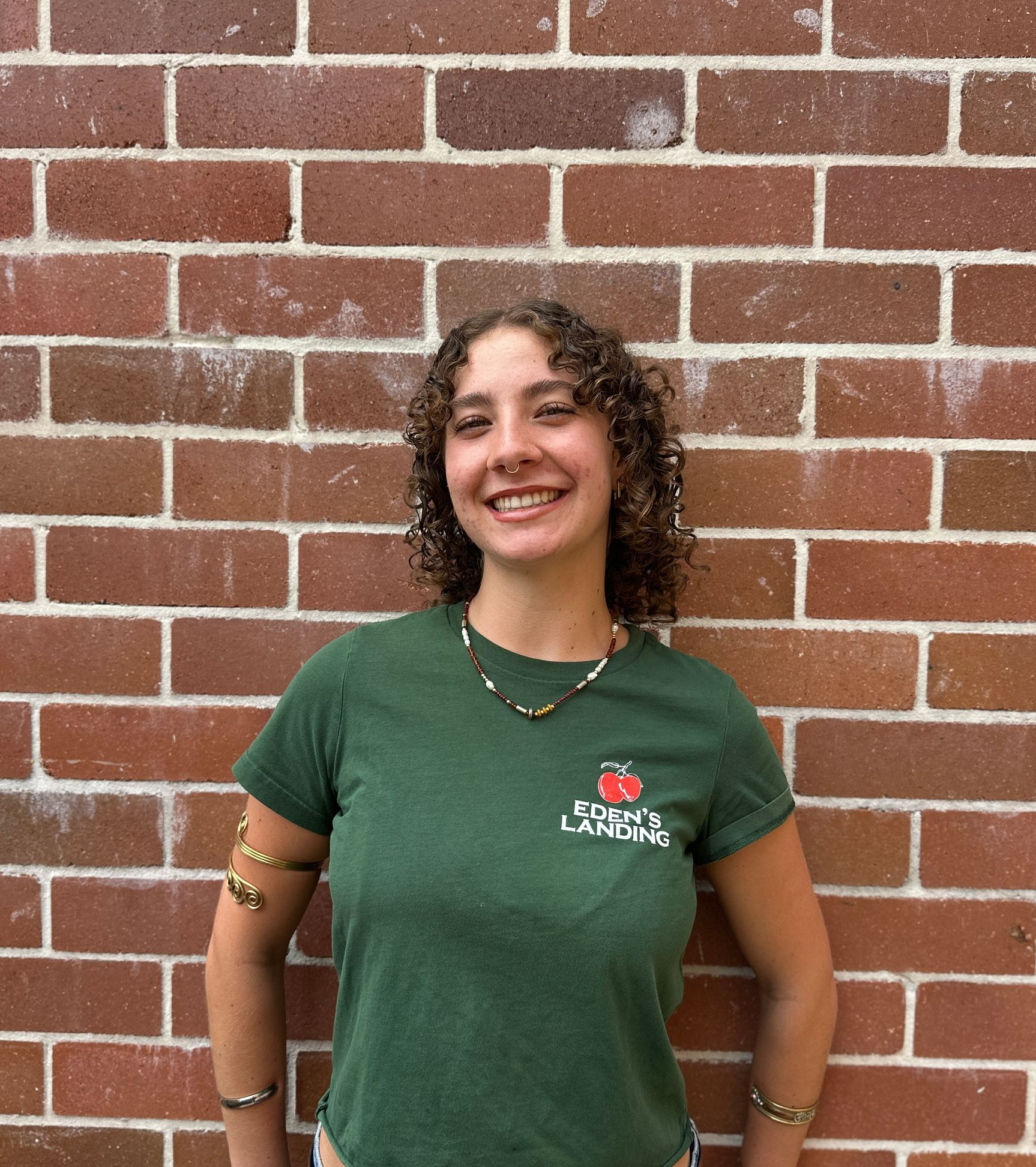 Woman in green shirt smiles in front of a brick wall, hands on hips, wearing jewelry.