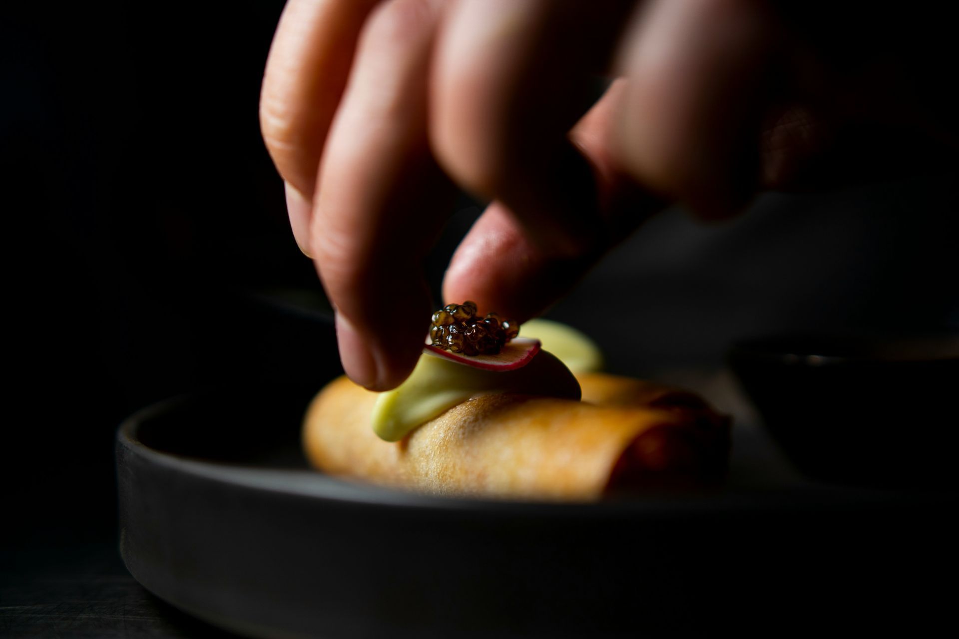 A chef's hand garnishes a small, golden spring roll with caviar and avocado on a dark plate.