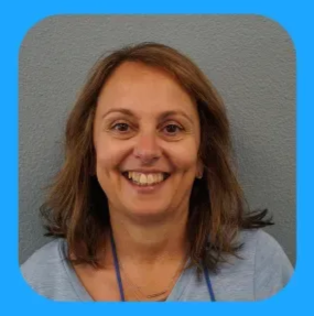 Woman smiling, wearing a light blue shirt and a blue lanyard, in front of a gray wall.