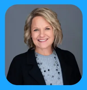 Woman with short blonde hair smiles, wearing a black blazer and gray necklace against a gray background.