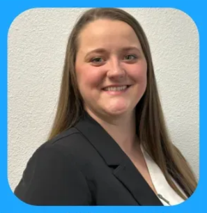 Woman in a black blazer and white blouse smiles, against a white wall and blue frame.