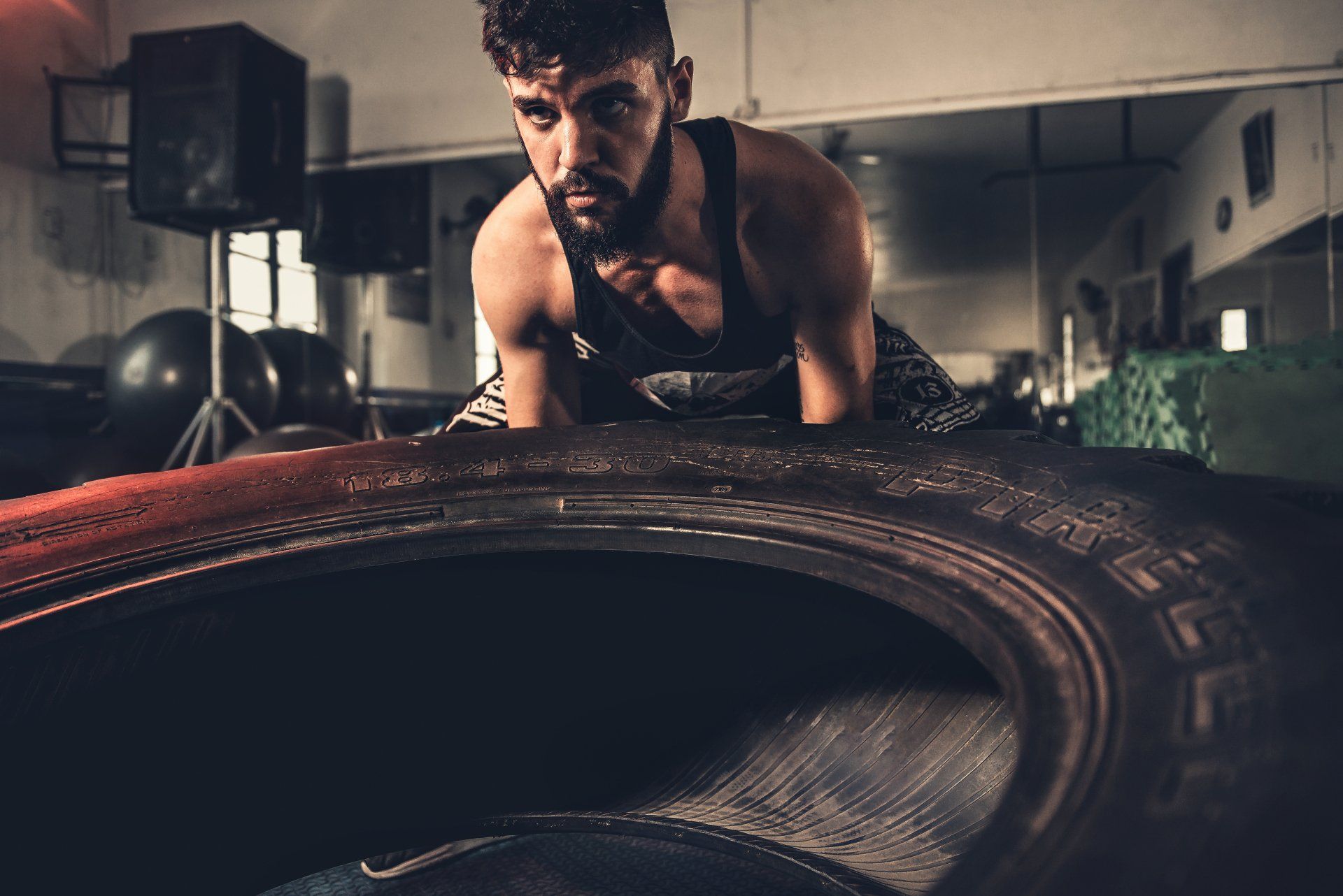 Man in a gym, pushing a large tire. Dark setting, intense expression.