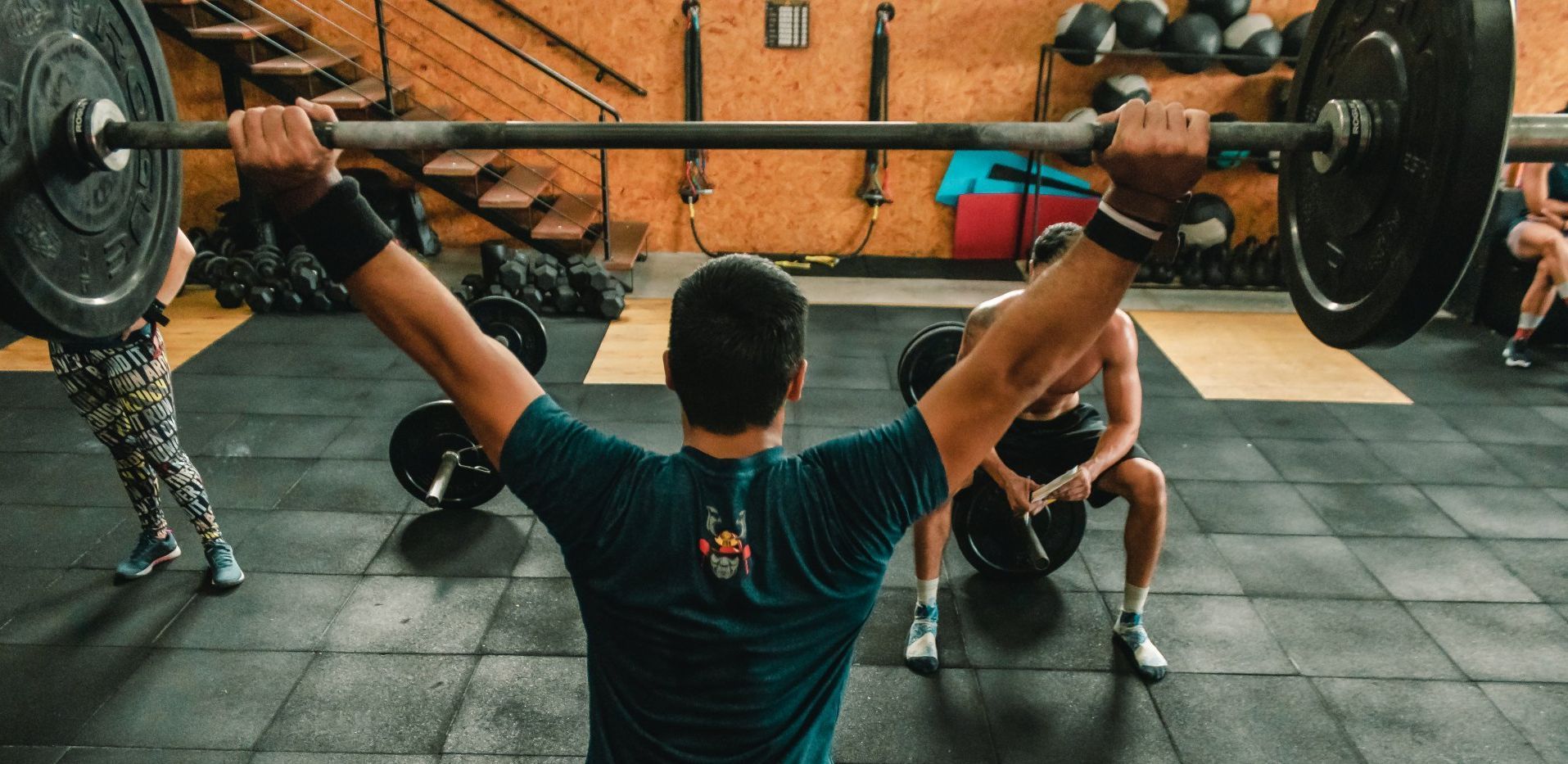 Person lifting a barbell overhead in a gym, others nearby.