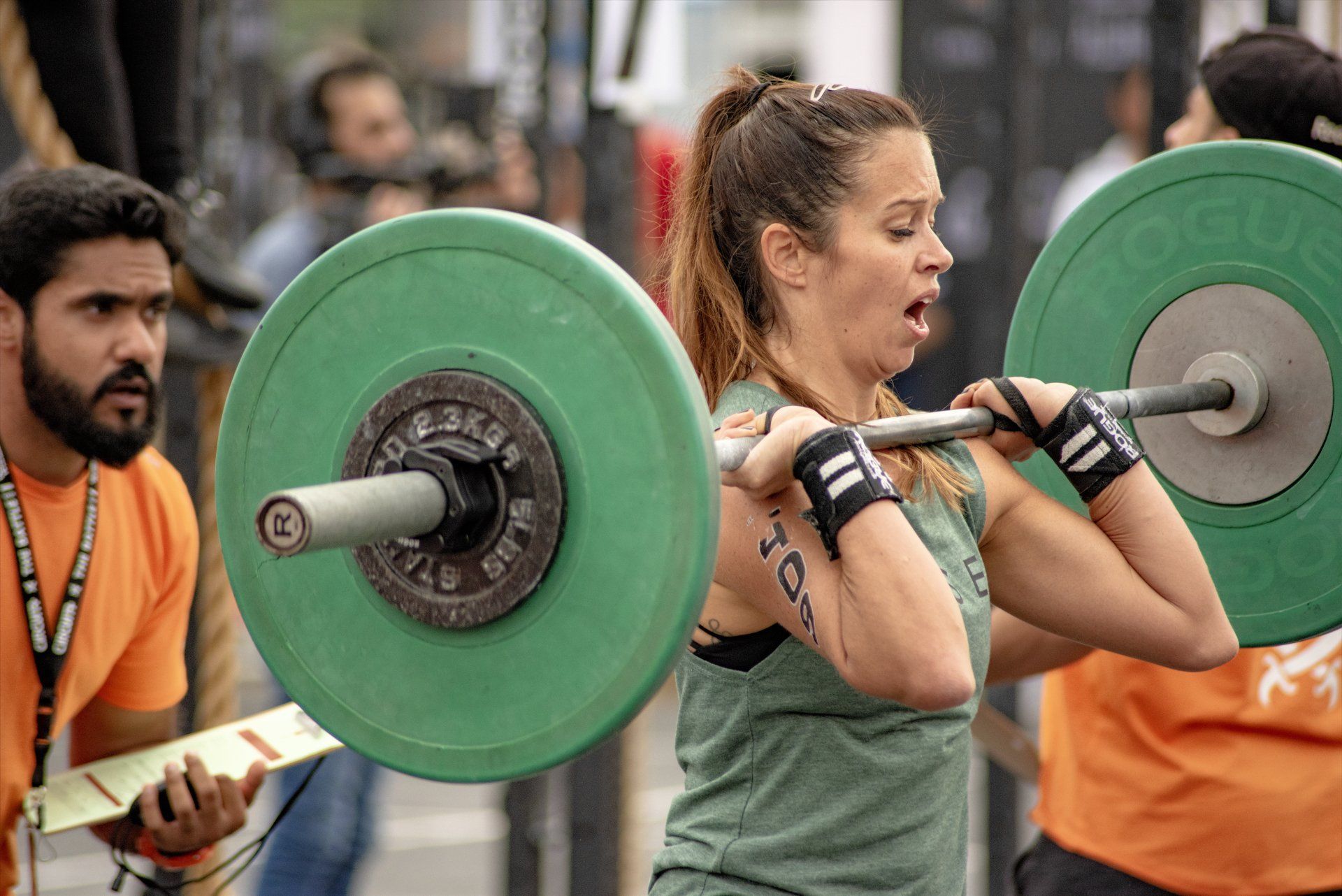 Woman in workout gear lifts a barbell during competition, sweating and grimacing. A judge watches.