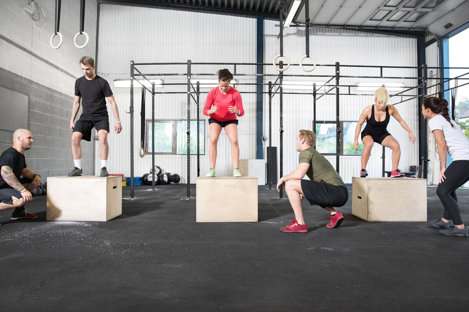 People doing box jumps at a gym; others watch. Black floor, wooden boxes, metal racks.