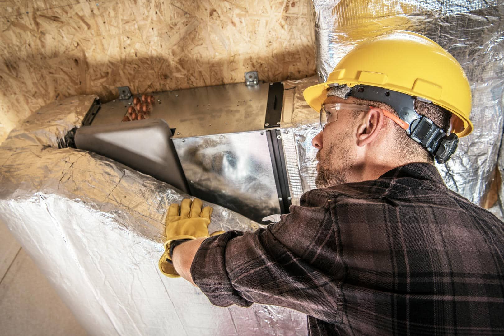Construction worker in a yellow hard hat installing ductwork in an attic, wearing gloves.