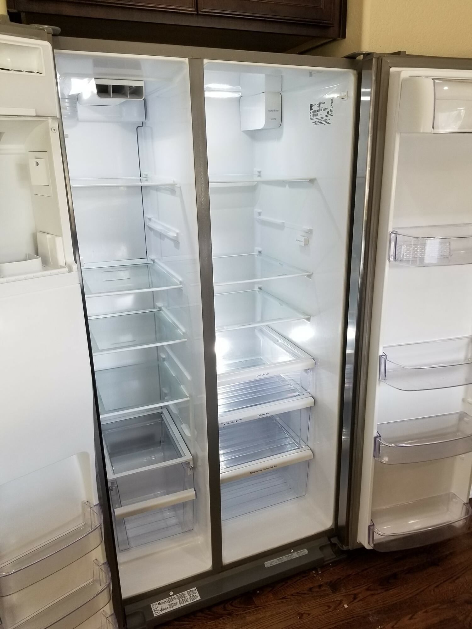 An empty refrigerator with the doors open is sitting on a wooden floor.