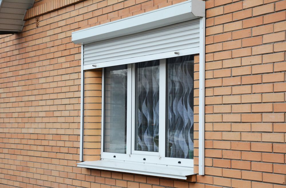 White roller shutter installed over a double-glazed window on a brick exterior wall for security