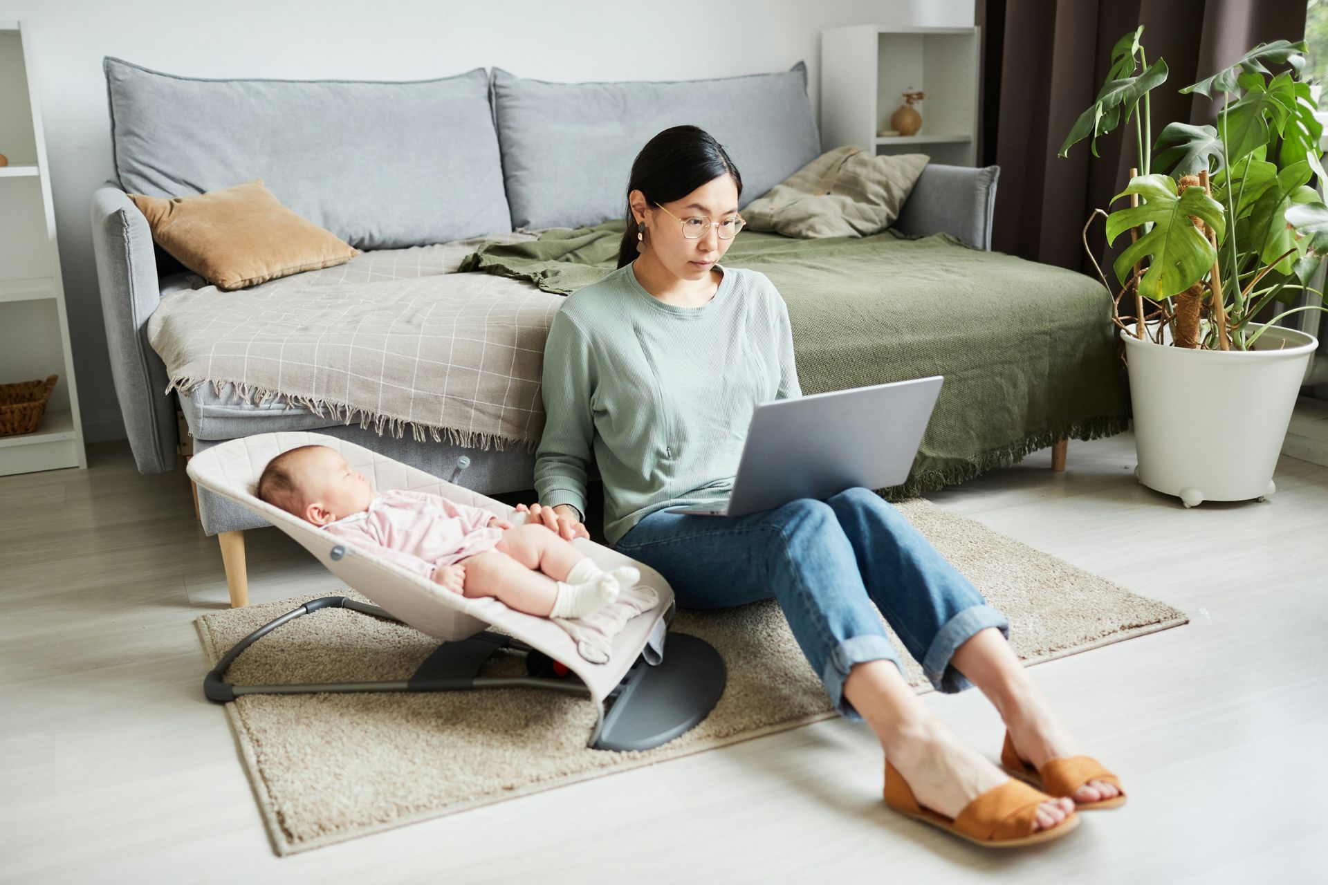 A woman is sitting on the floor with a baby in a bouncer and using a laptop.