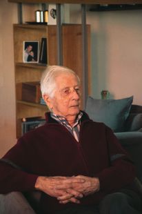 An elderly man is sitting on a couch with his hands folded.