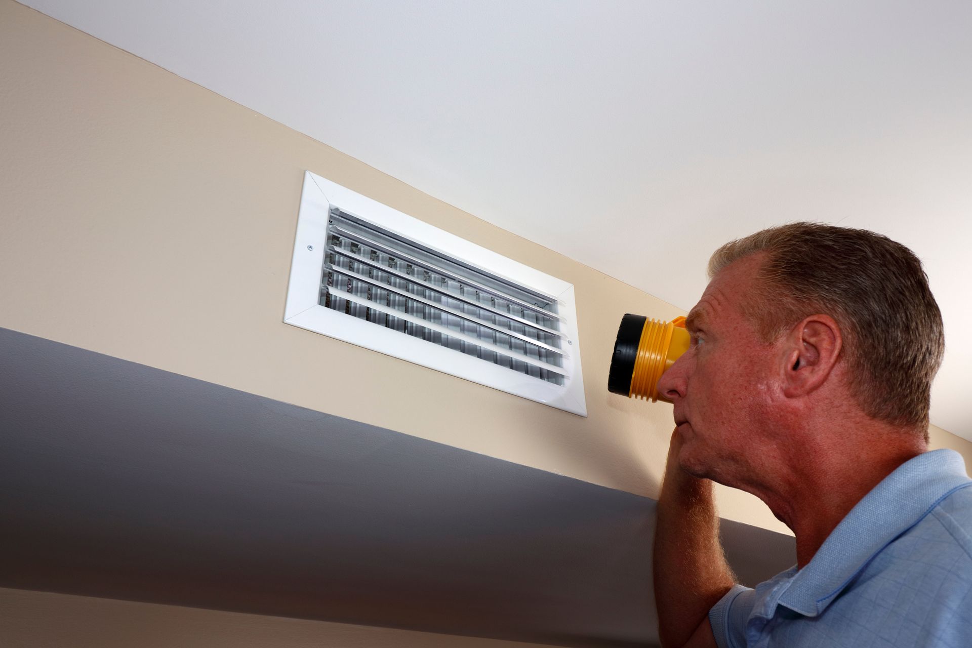 Man inspecting an air vent with a flashlight on a beige wall