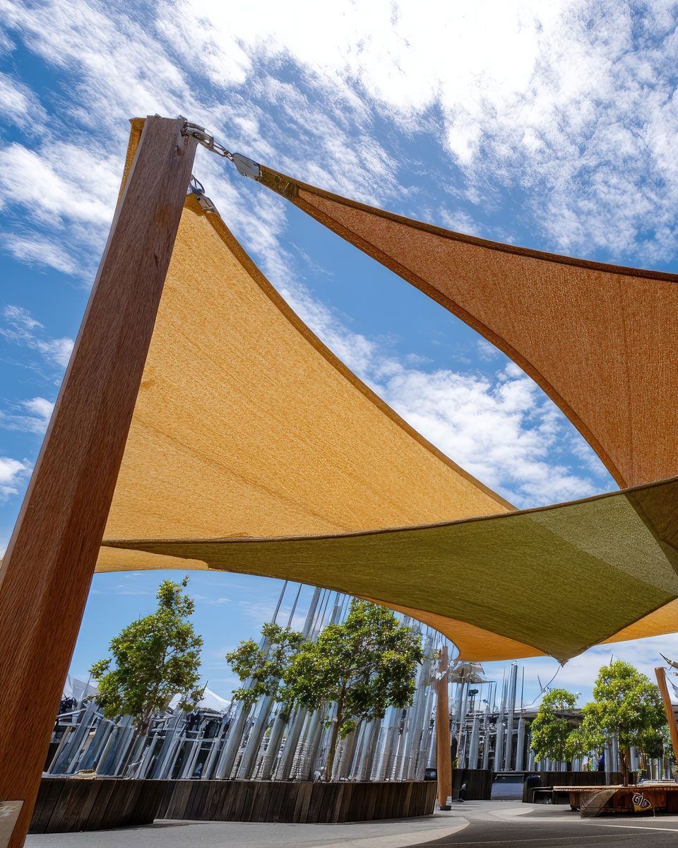 Triangular shade sails over a park. Two sails are beige and olive green, against a blue sky.