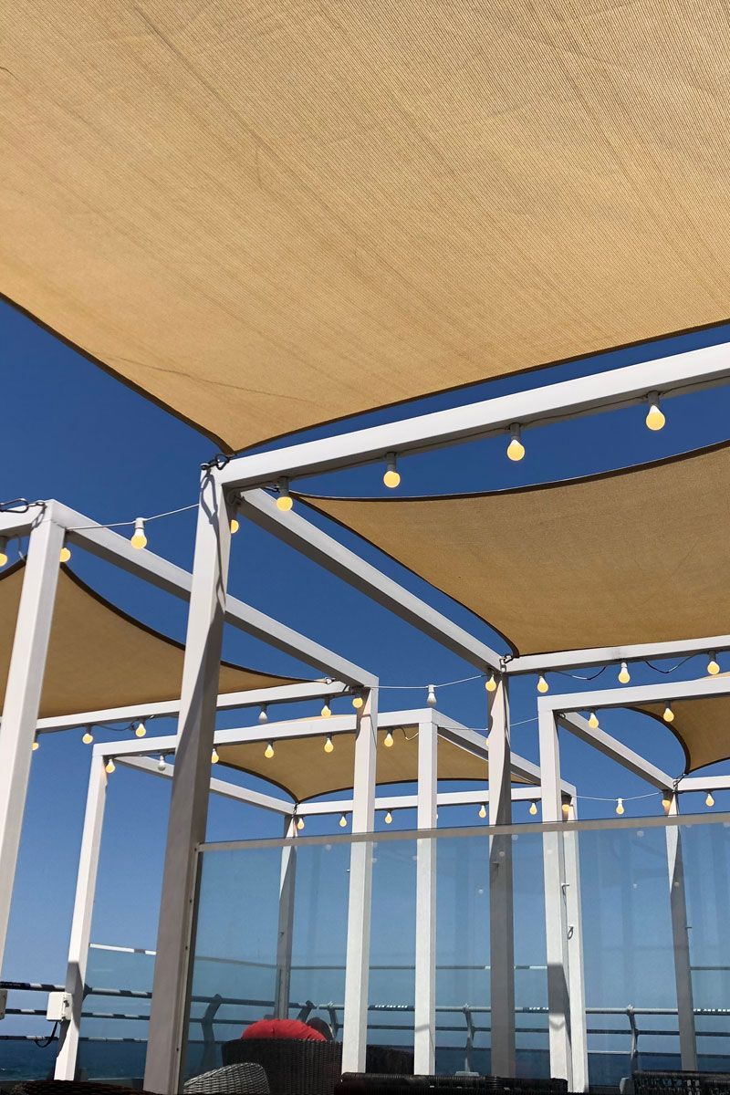 Outdoor seating area with white frames, tan shade cloths, and string lights, against a blue sky.