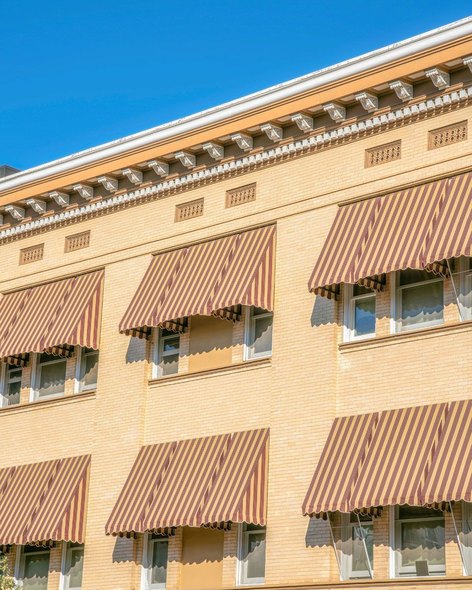 Tan brick building with brown striped awnings over windows against a blue sky.