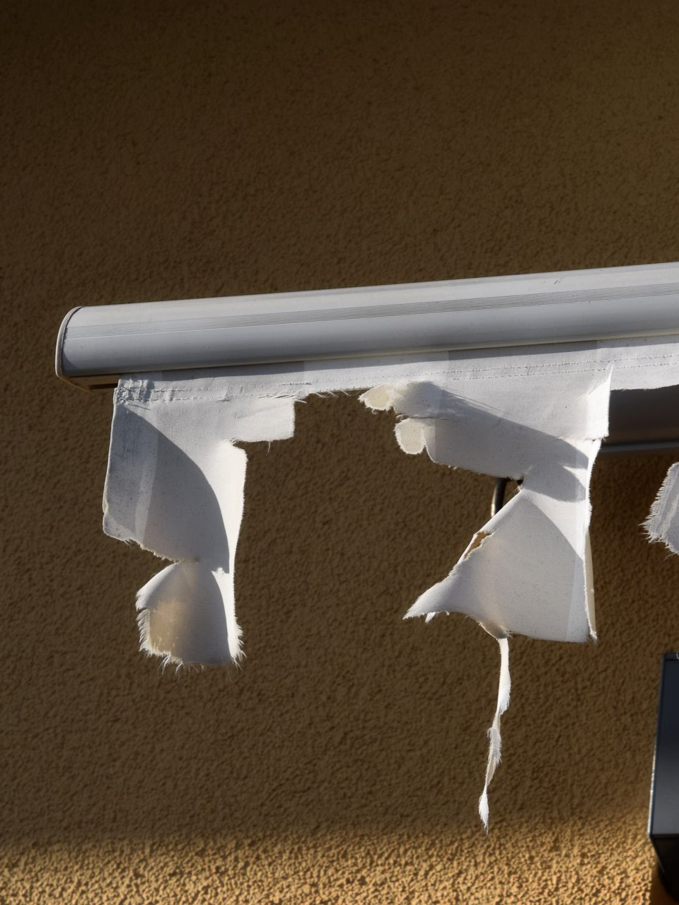 Torn, white awning fabric hanging from a white metal frame, against a tan wall.