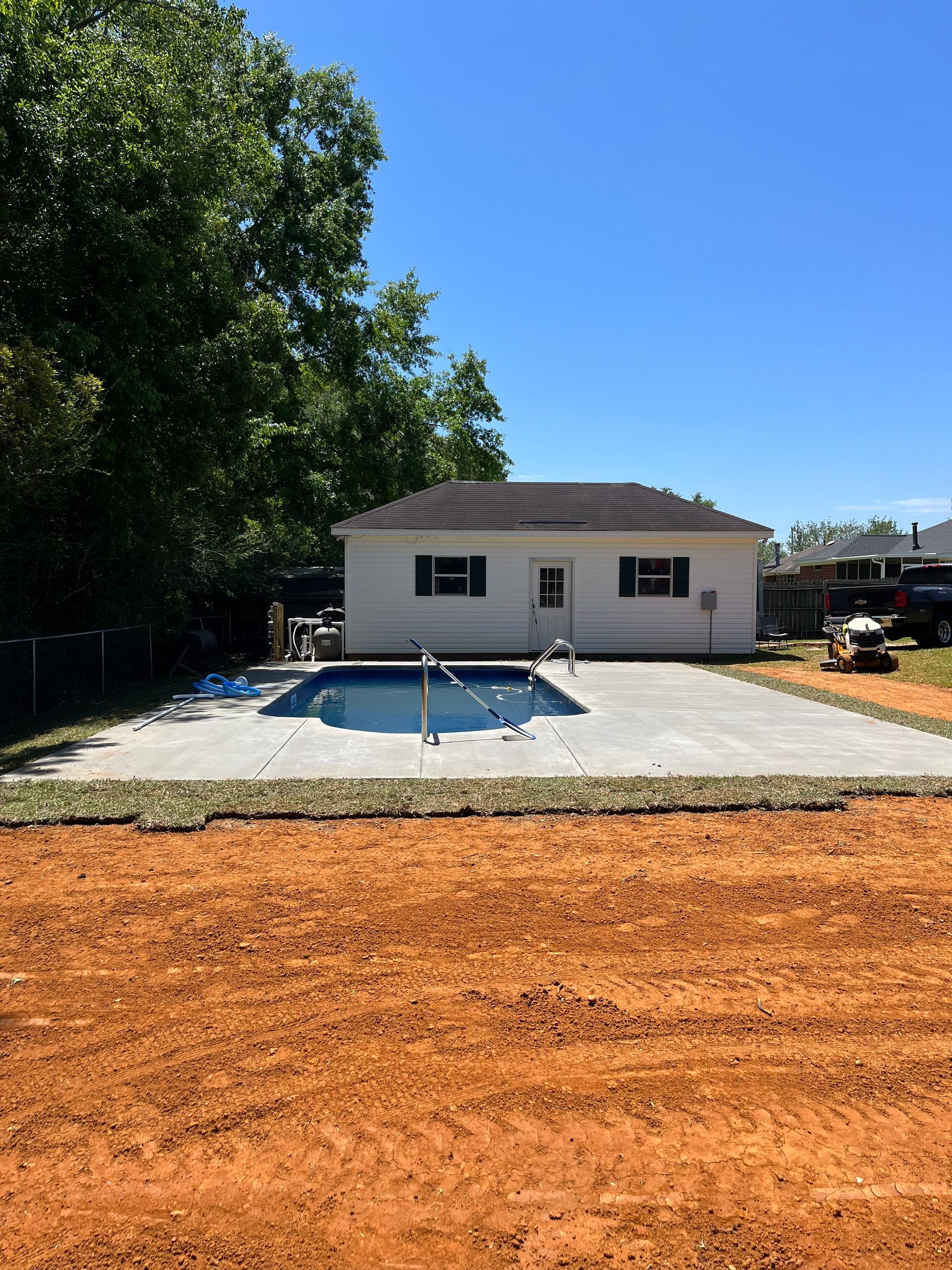 White house with in-ground pool on concrete patio; dirt foreground, trees in background.