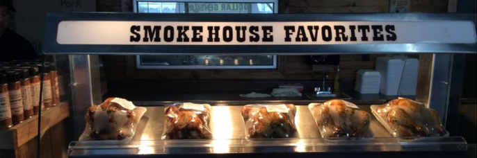 A heated food display case labeled "SMOKEHOUSE FAVORITES" holding several whole roasted chickens in plastic packaging.