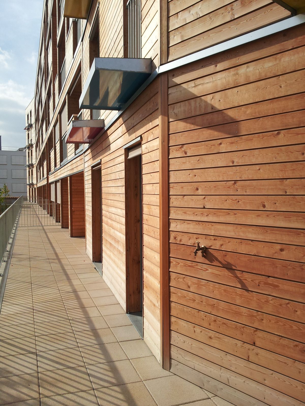 A sunlit exterior wooden balcony of an apartment building with several doorways, lined with tiled flooring and a rail.