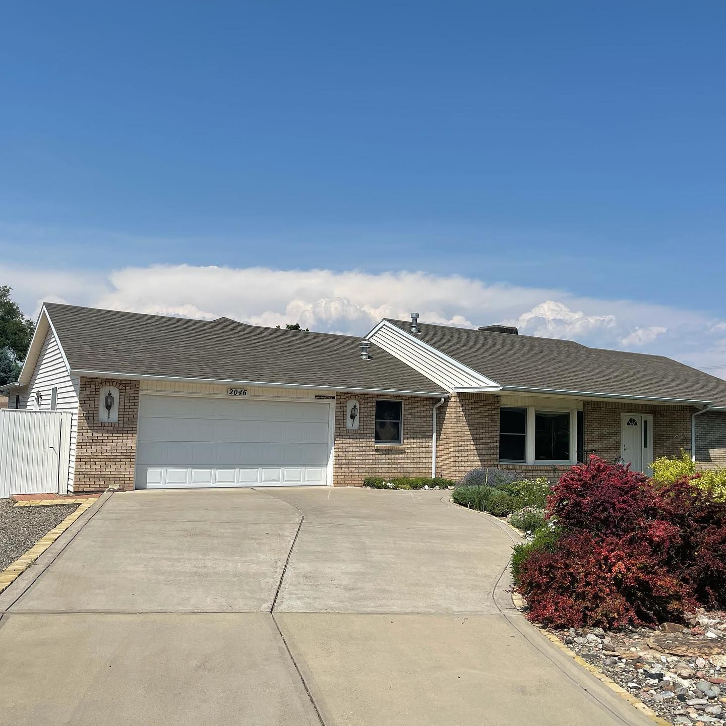 Ranch-style house with gray roof, beige siding, white garage door, concrete driveway, and landscaping under a blue sky.