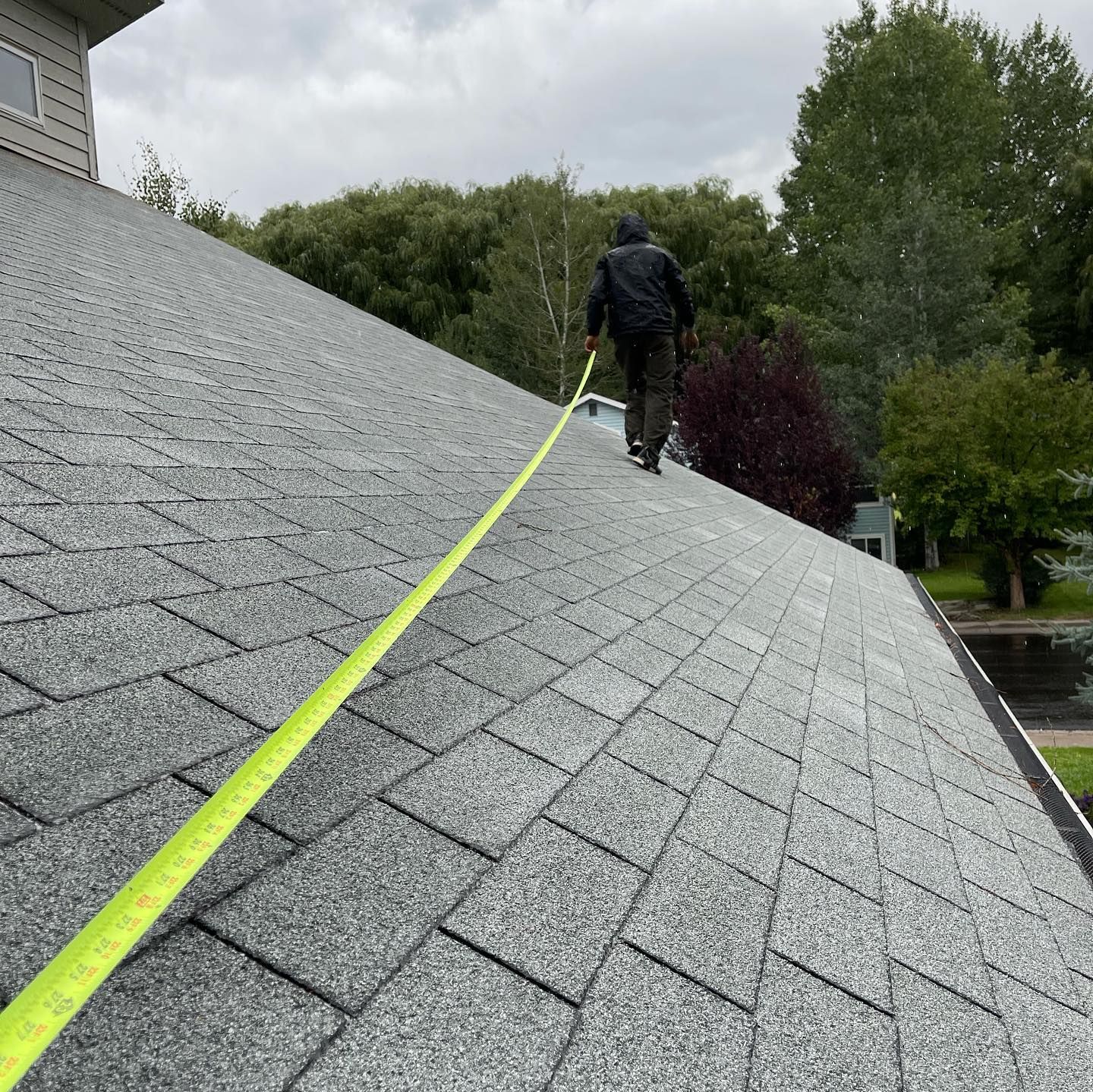Person on a gray shingle roof measuring with a yellow tape. Trees in the background, overcast sky.