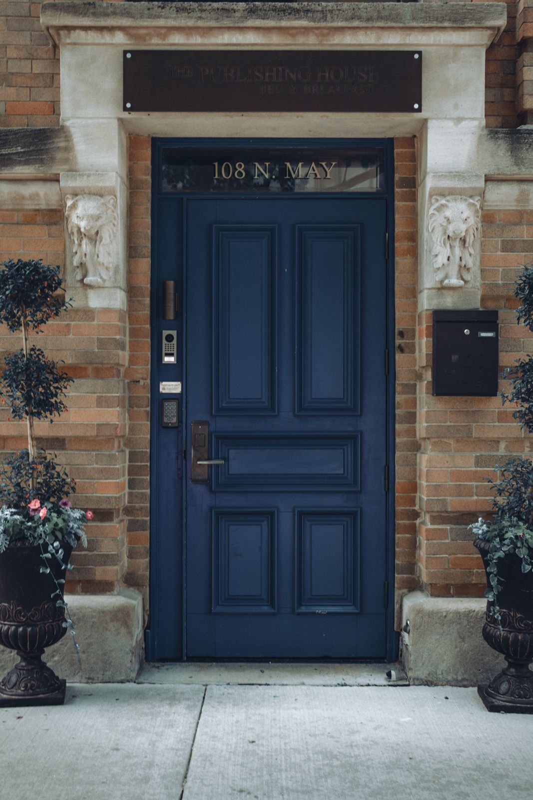 A dark blue front door with a transom window, set in a brick building frame with stone details and flanking potted plants.