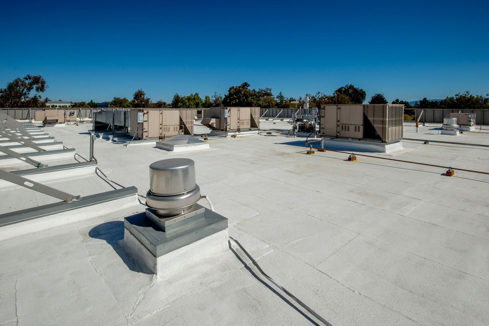 A flat white commercial roof under a clear blue sky, featuring several HVAC units and a metallic ventilation fan.