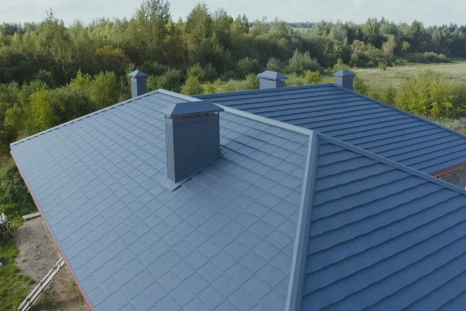 Blue metal roof of a house with chimneys, set against a backdrop of green trees.