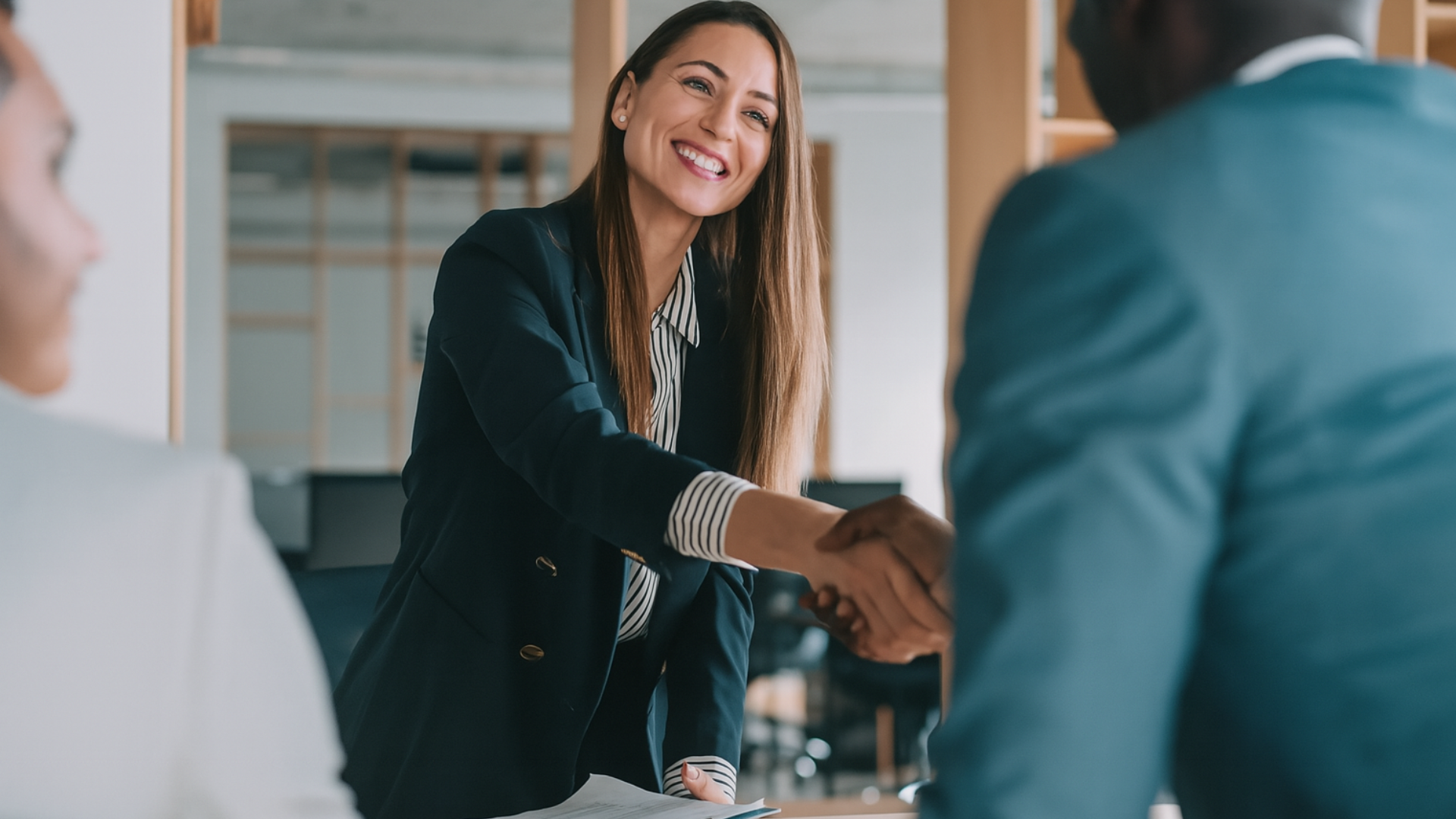 Woman in navy blazer shakes hands with a man in a suit, smiling in an office setting. Another person is seated nearby.