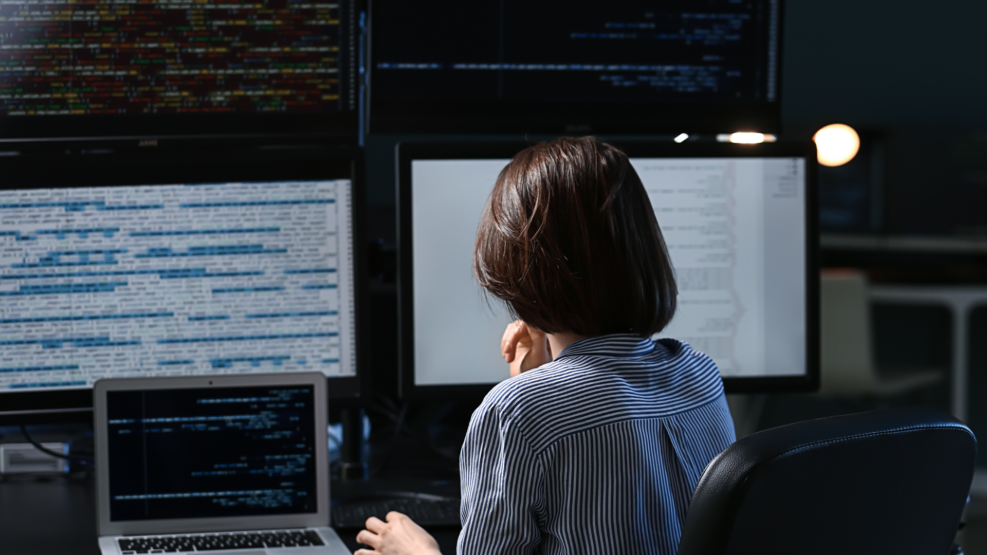 A woman with short hair sits at a desk, facing multiple computer screens displaying code.