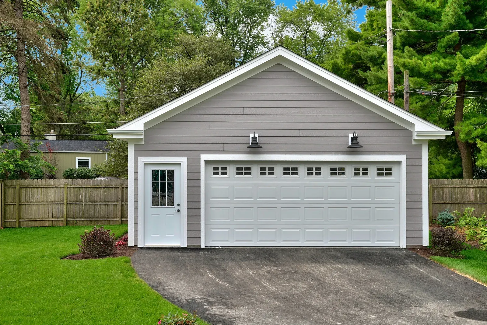 Gray garage with white door and garage door, asphalt driveway, green lawn, wooden fence.