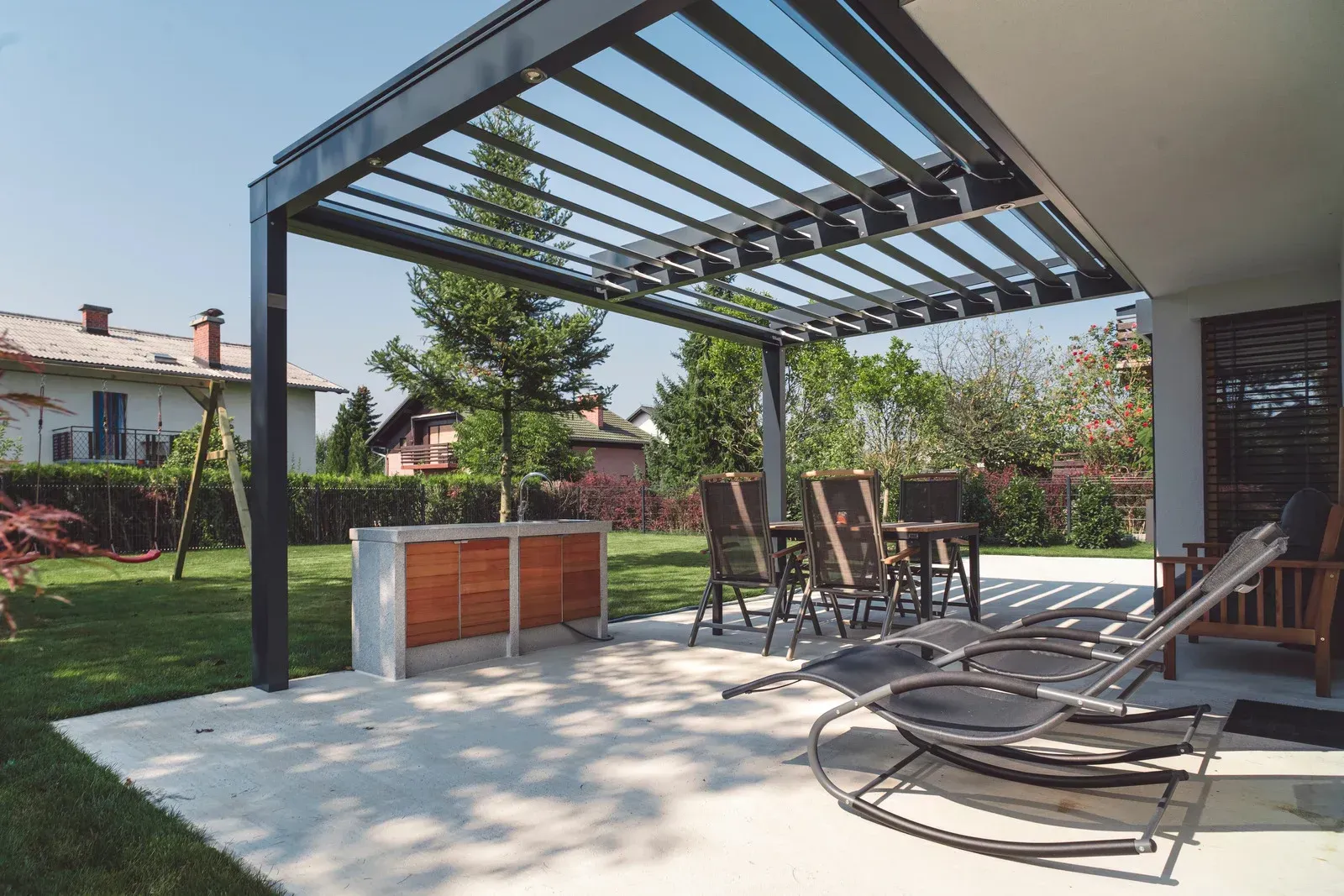 Patio with pergola, dining set, and lounge chairs, with a yard and house in background.