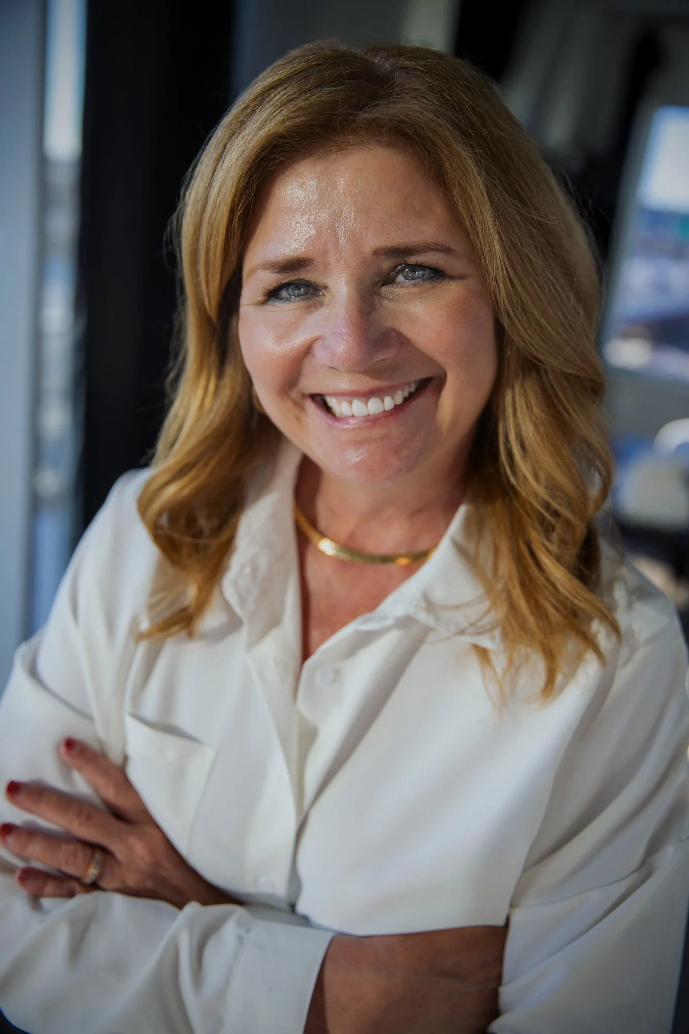 Smiling woman in a white blouse with arms crossed in a softly lit indoor setting