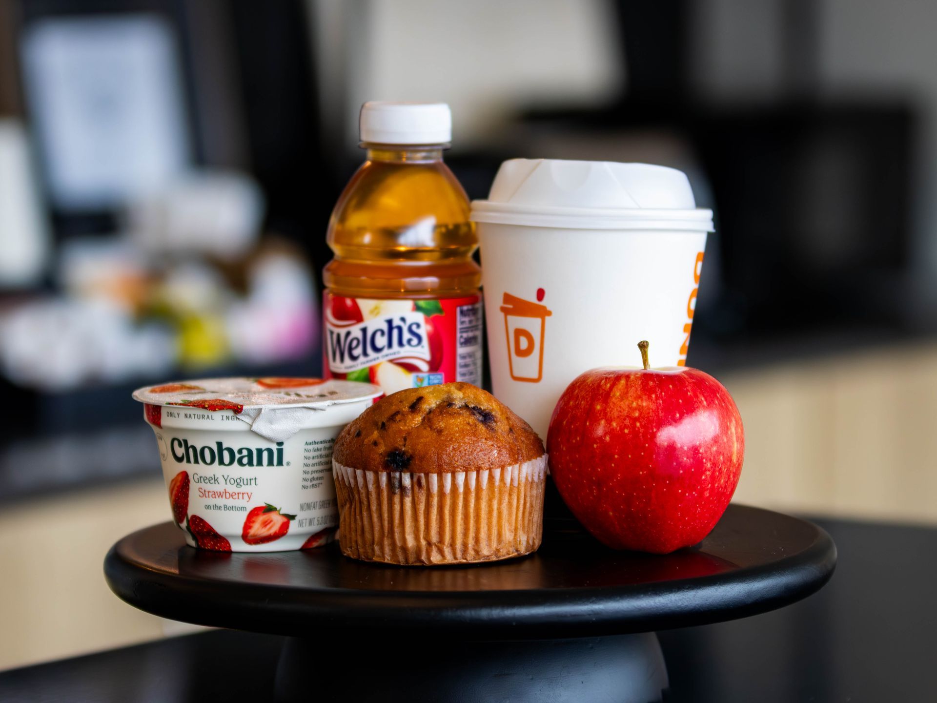 Breakfast spread on a black table: yogurt, muffin, apple, coffee cup, and a juice bottle.
