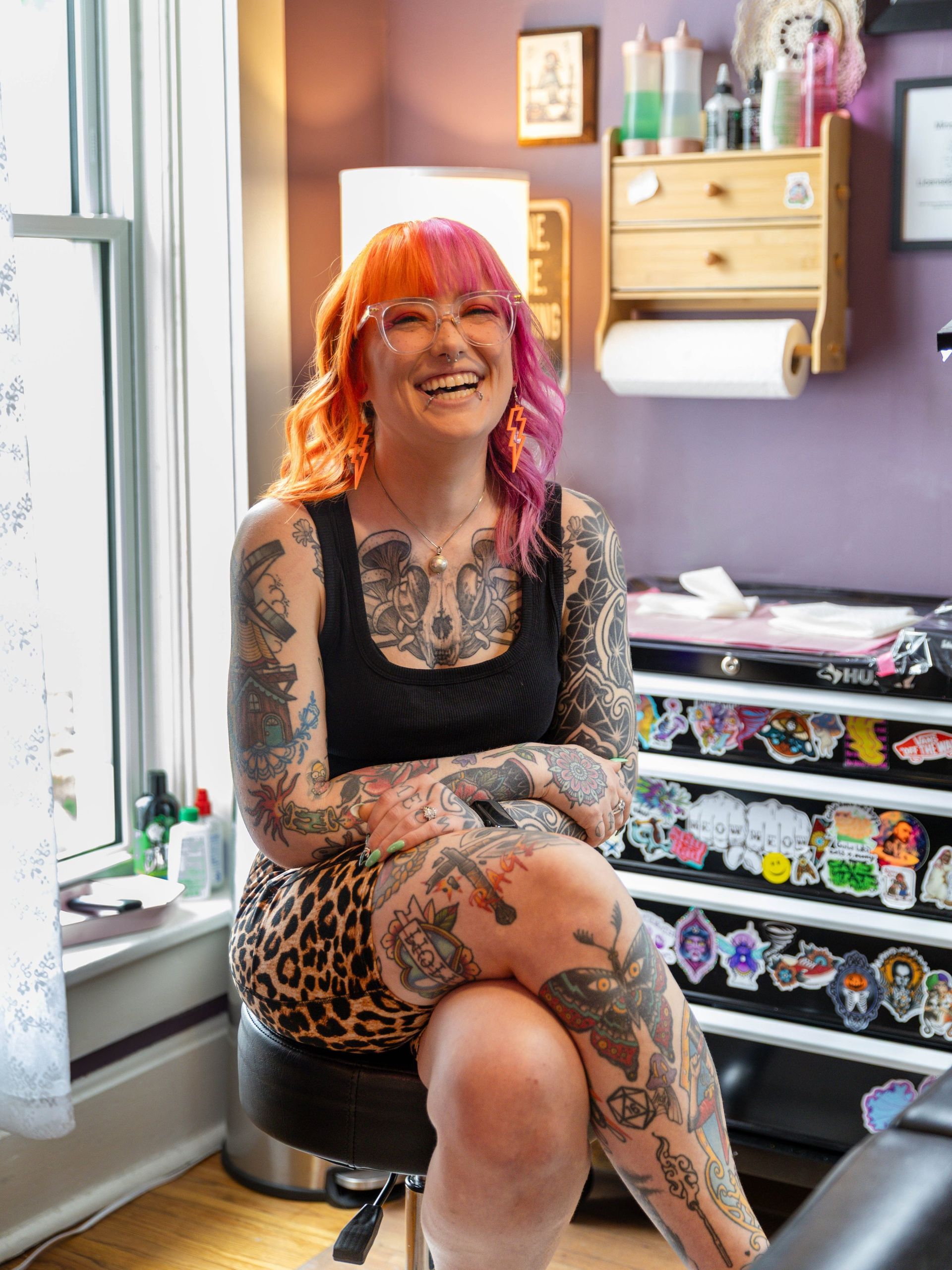 Person with orange and pink hair sitting on a stool in a purple bedroom, smiling with tattooed arms and legs