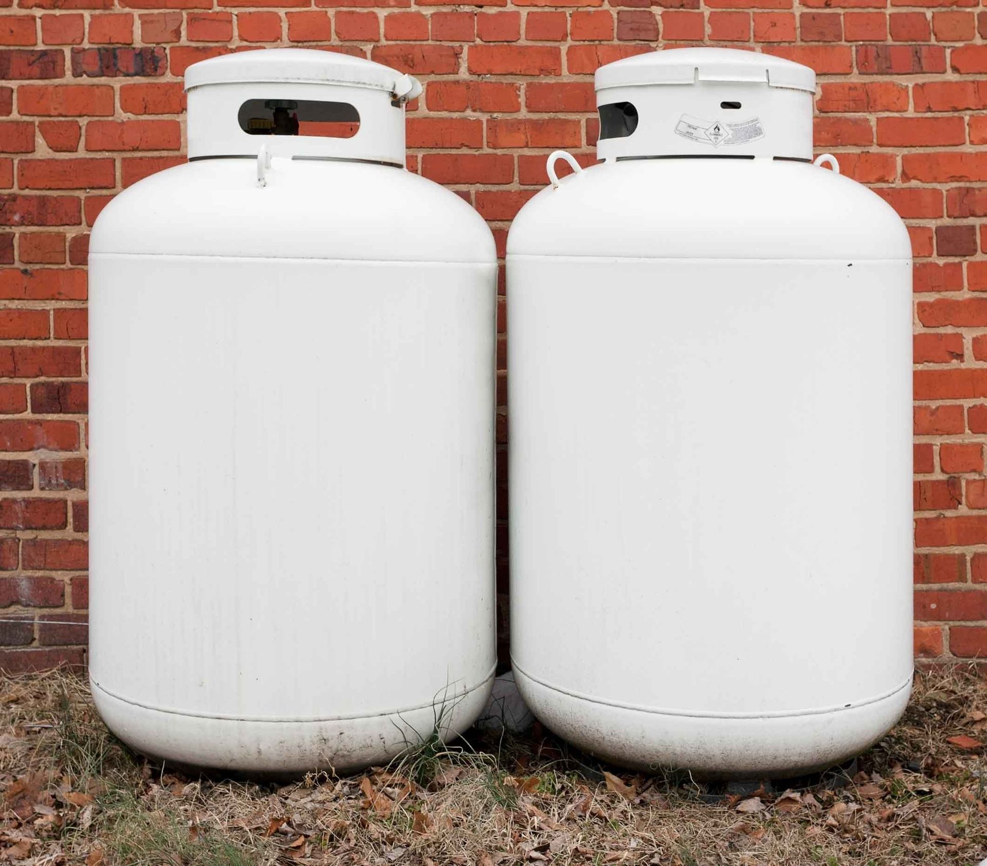 Two white propane tanks standing side by side against a red brick wall.