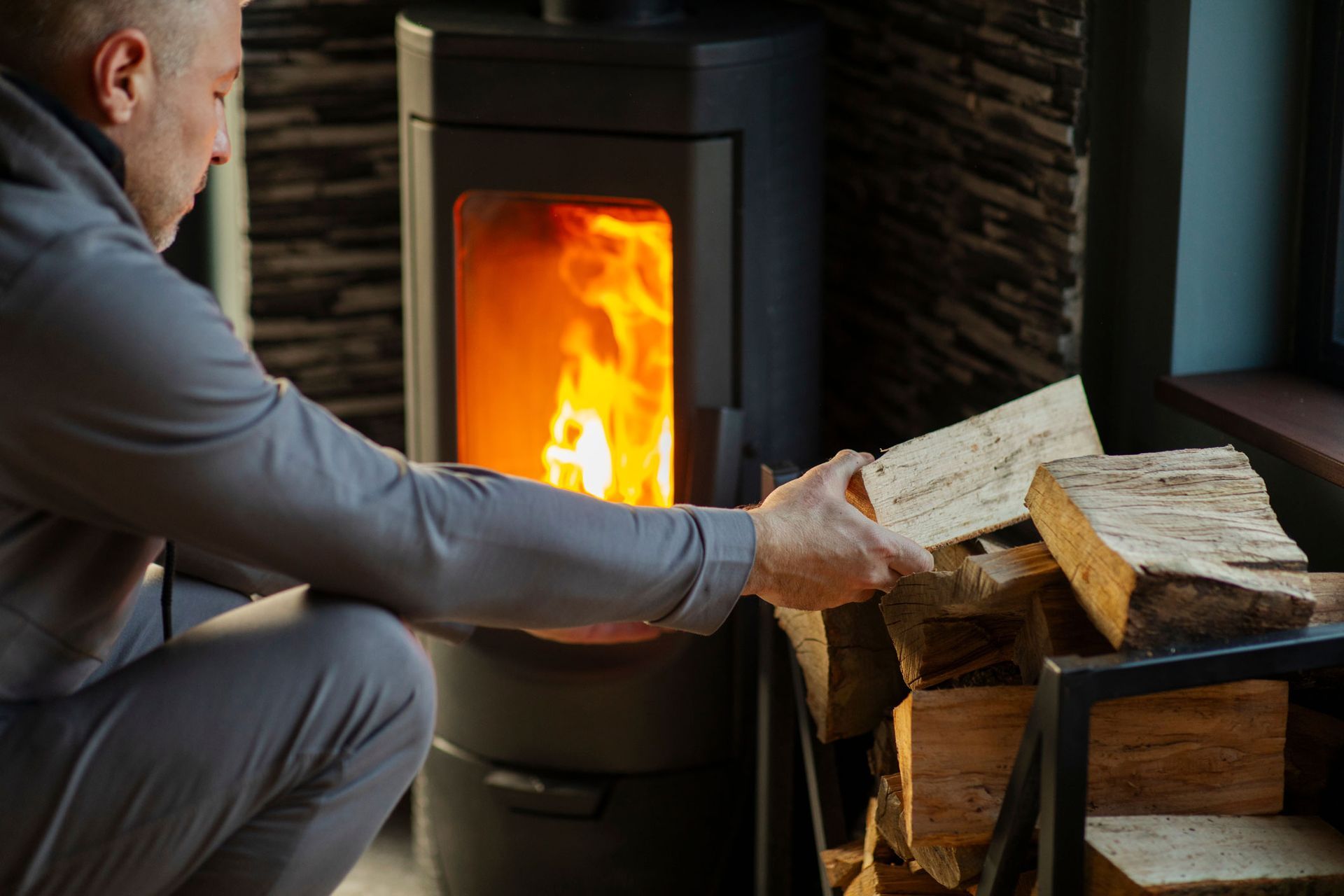 Person adding logs to a lit wood stove beside a stack of firewood