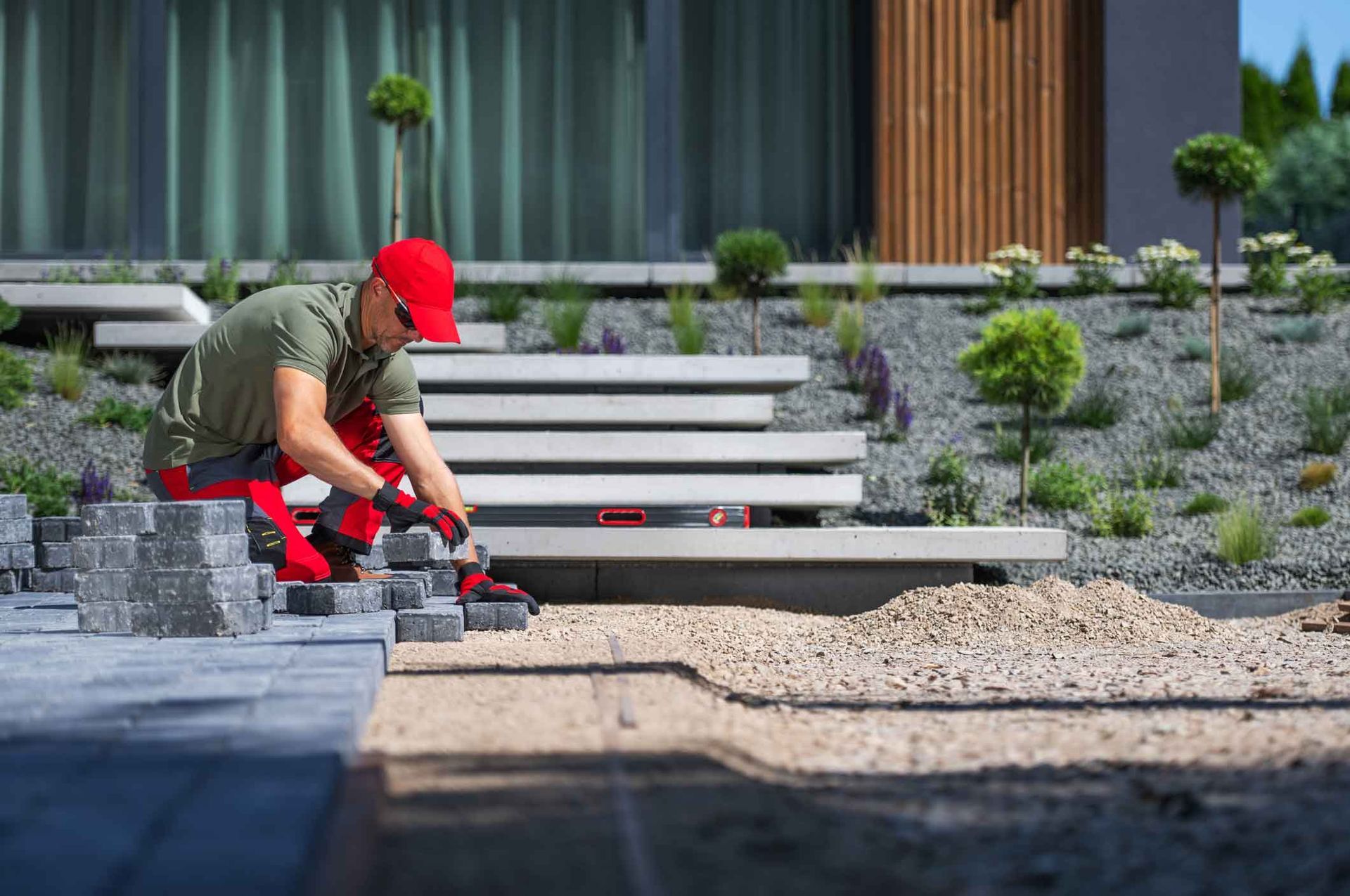 Worker in red cap kneeling by a stone walkway, placing paving stones in a landscaped yard.