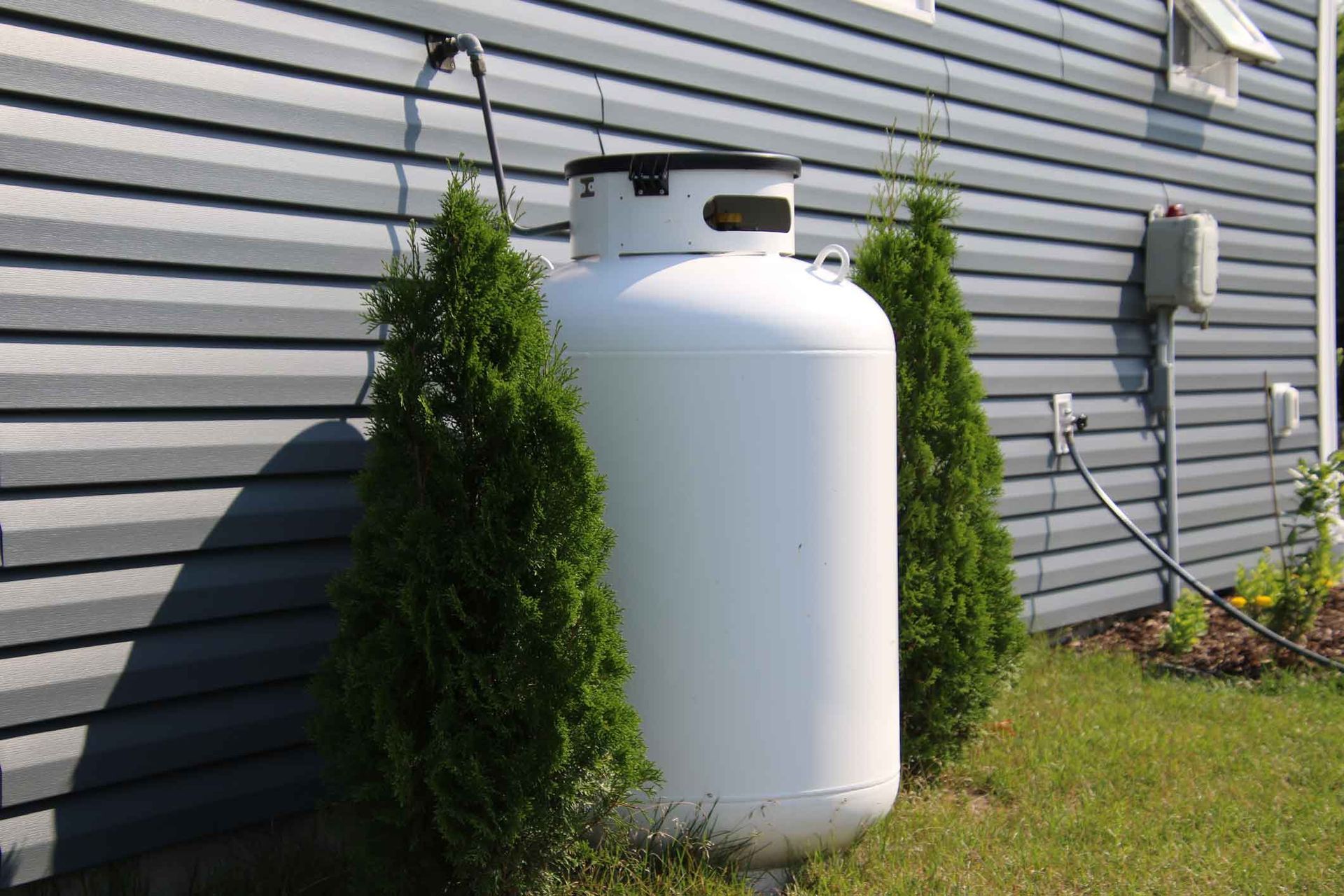 White propane tank beside a gray house wall with green bushes and grass