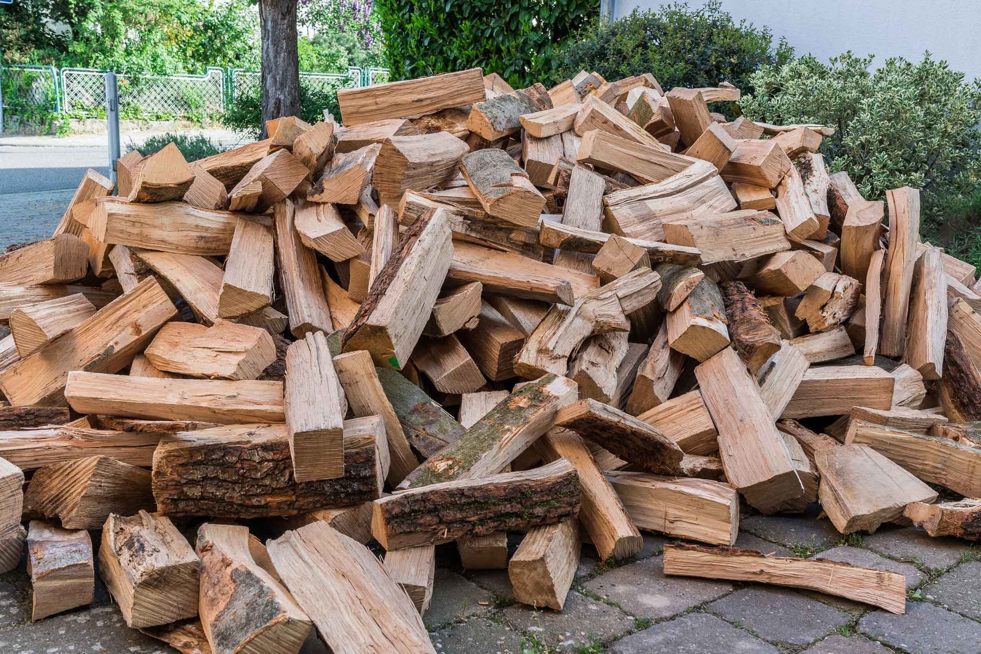 Pile of split firewood stacked outdoors on a paved area, with trees and bushes in the background