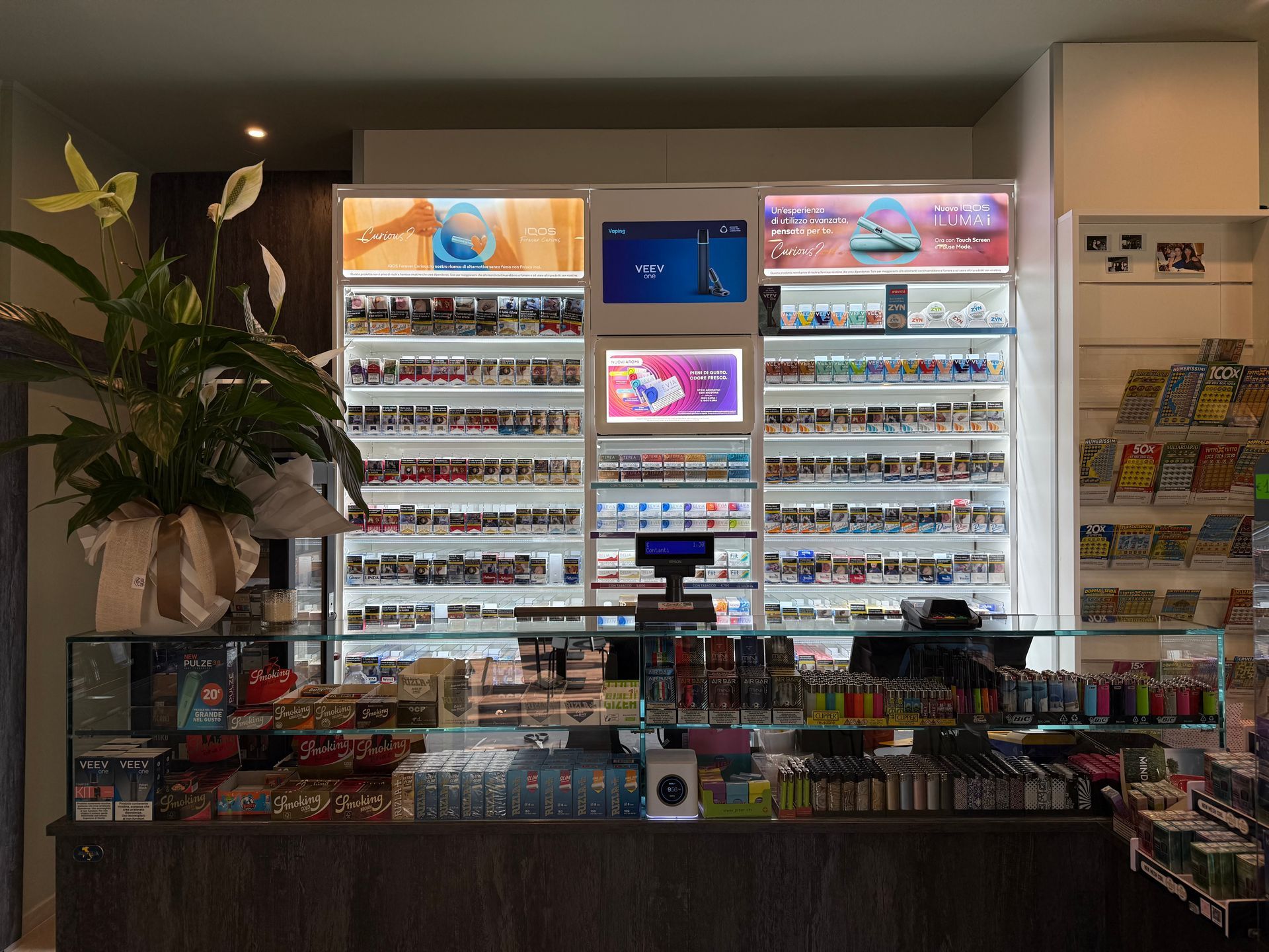 Il bancone di un minimarket con espositori illuminati di vari prodotti, tra cui tabacco e sigarette elettroniche.